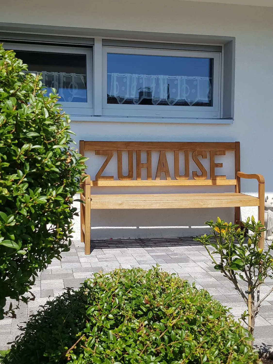 Mi Casa-Dein zu Hause - Familien- und Seniorenbetreuung - Eine hölzerne Gartenbank mit dem in die Rückenlehne eingravierten deutschen Wort „ZUHAUSE“ steht auf einer Steinterrasse vor einem weißen Haus mit großem Fenster. Grüne Büsche rahmen die Szene im Vordergrund und links ein.